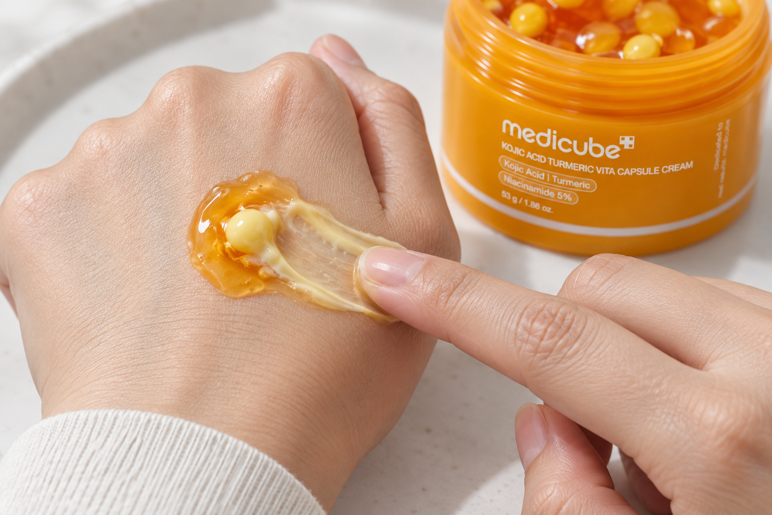 Un dedo integra una cápsula amarilla con el gel anaranjado de la crema Medicube sobre el dorso de una mano